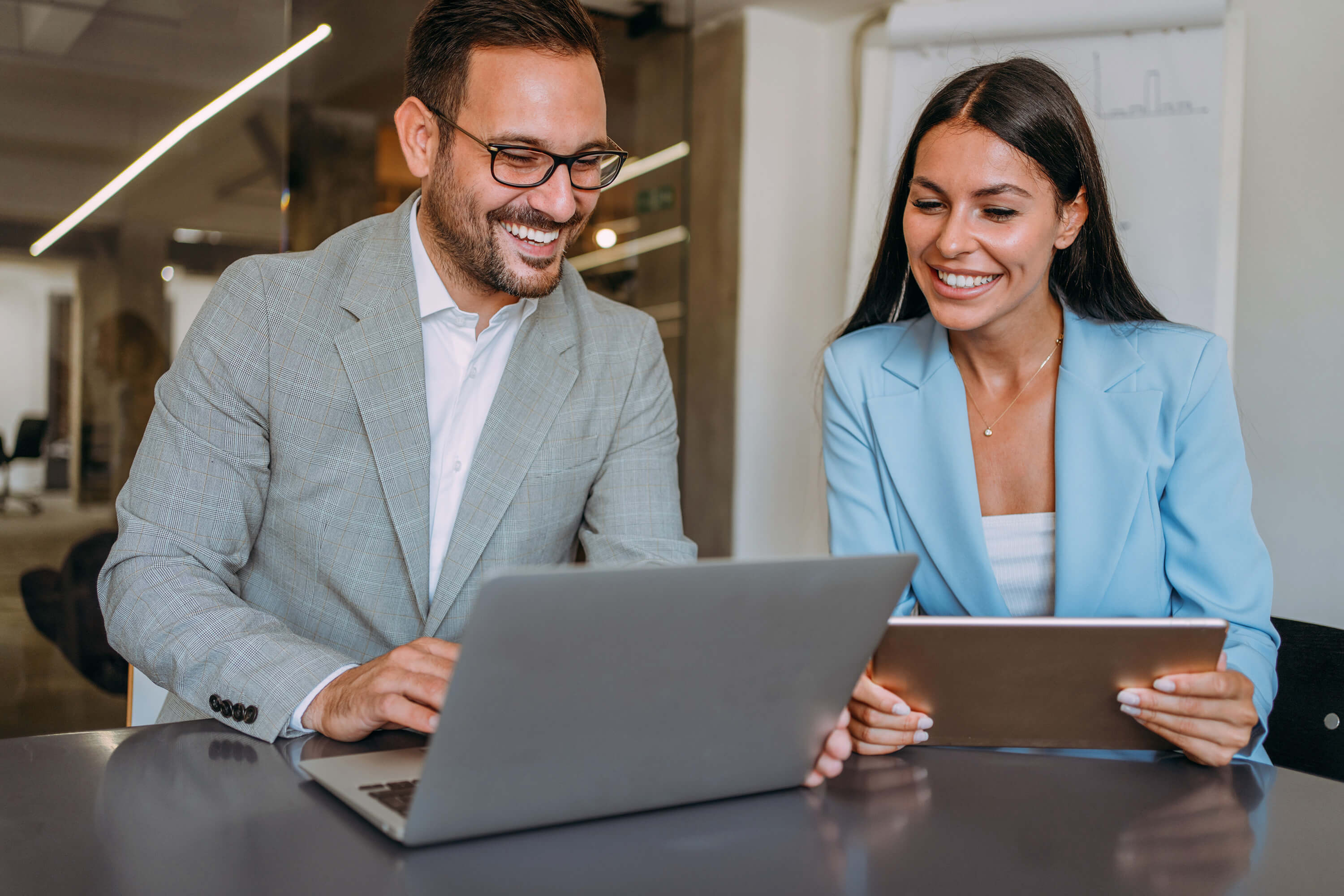 A professional man and woman sit at a table in their office and smile as they compare information on his laptop and her tablet.