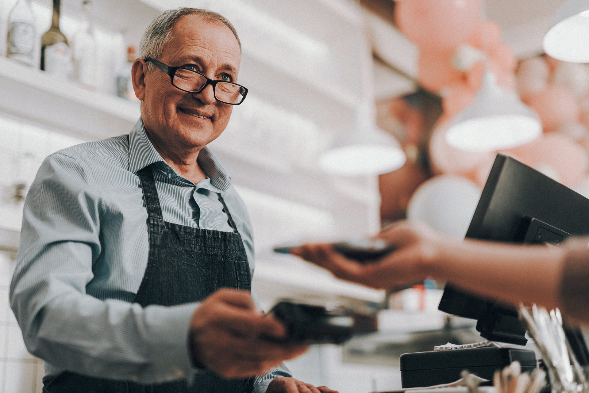 An older man smiles as helps a customer at a cash register.