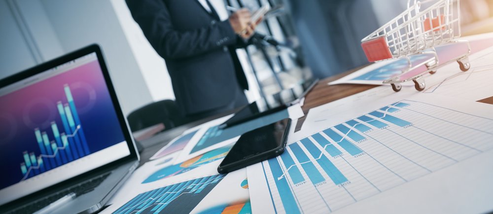 office desk with investment charts displayed and male in suit standing in background.