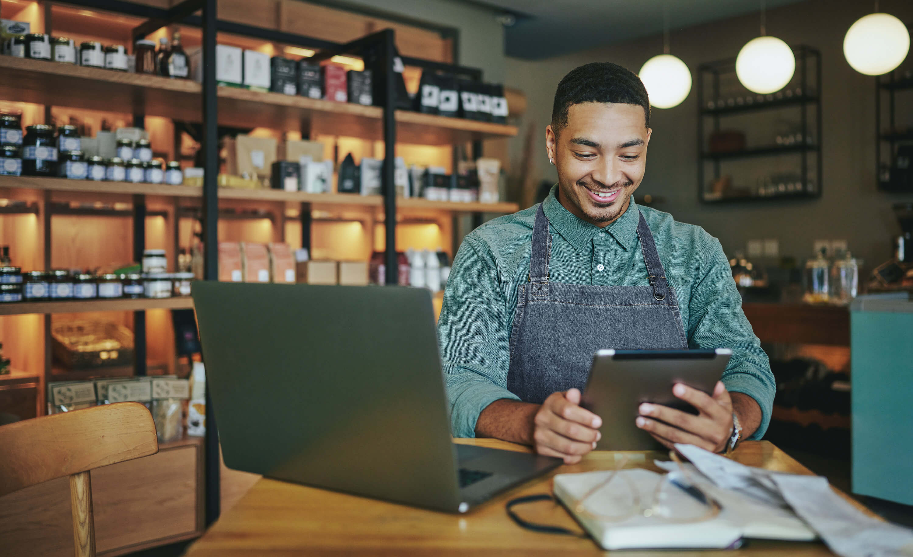 A black male small business owner wearing an apron stands at a table in his retail store and smiles as he looks down at his tablet.