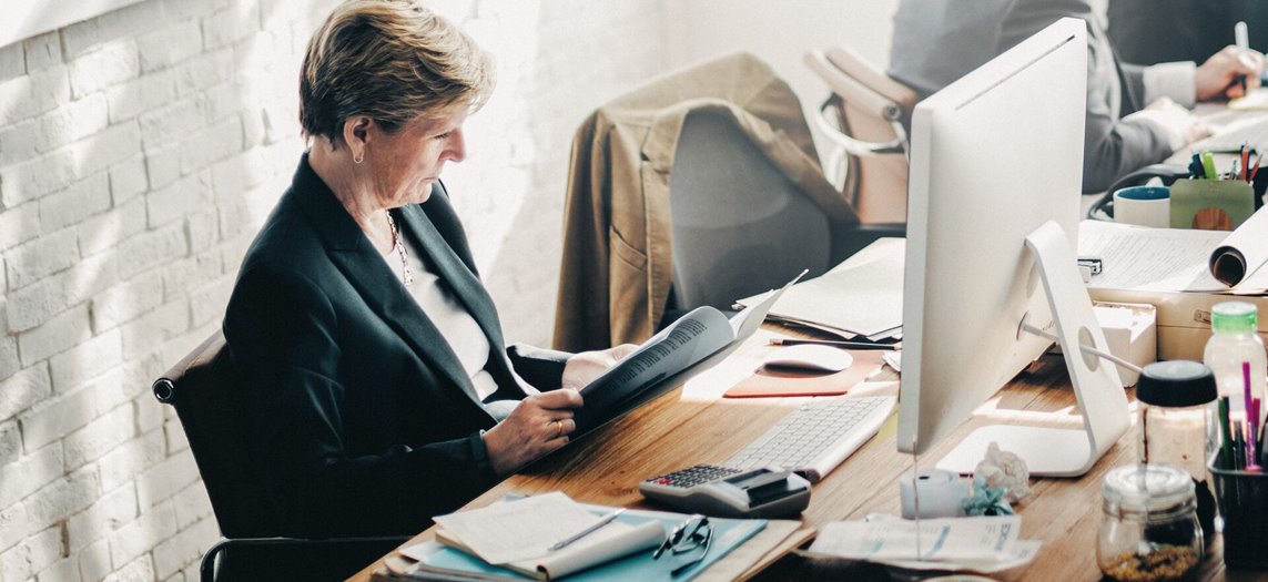 A woman in her fifties is wearing a suit and reading a document while sitting at a crowded desk in an industrial community office space.