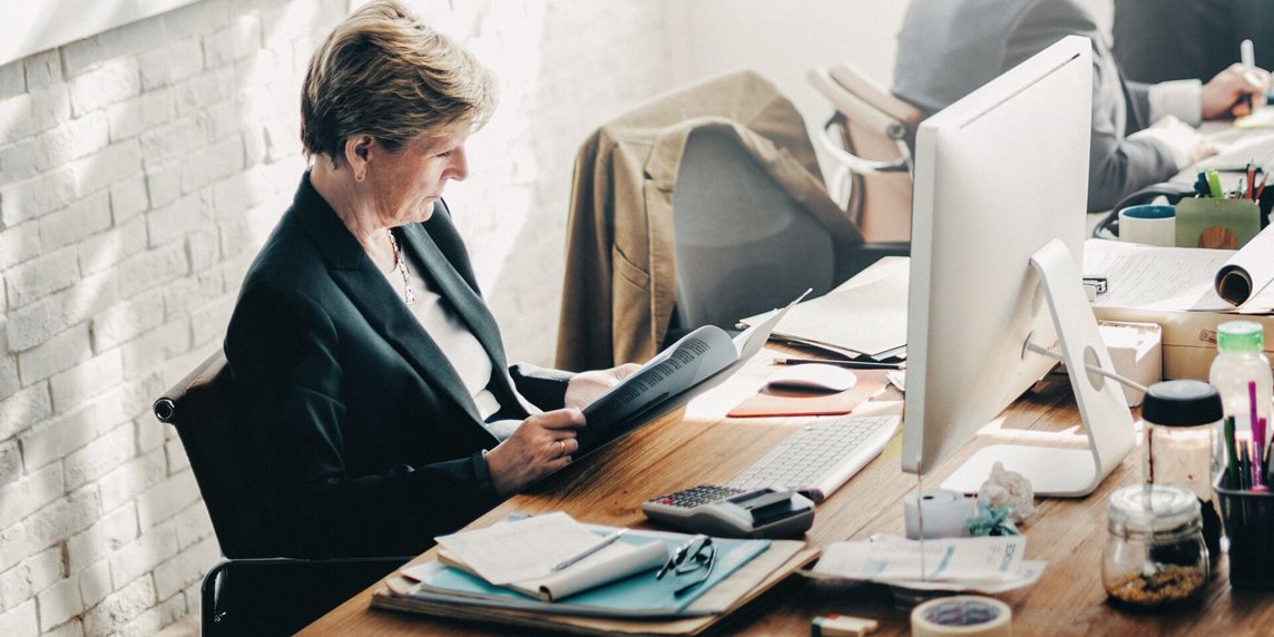 A woman in her fifties is wearing a suit and reading a document while sitting at a crowded desk in an industrial community office space.