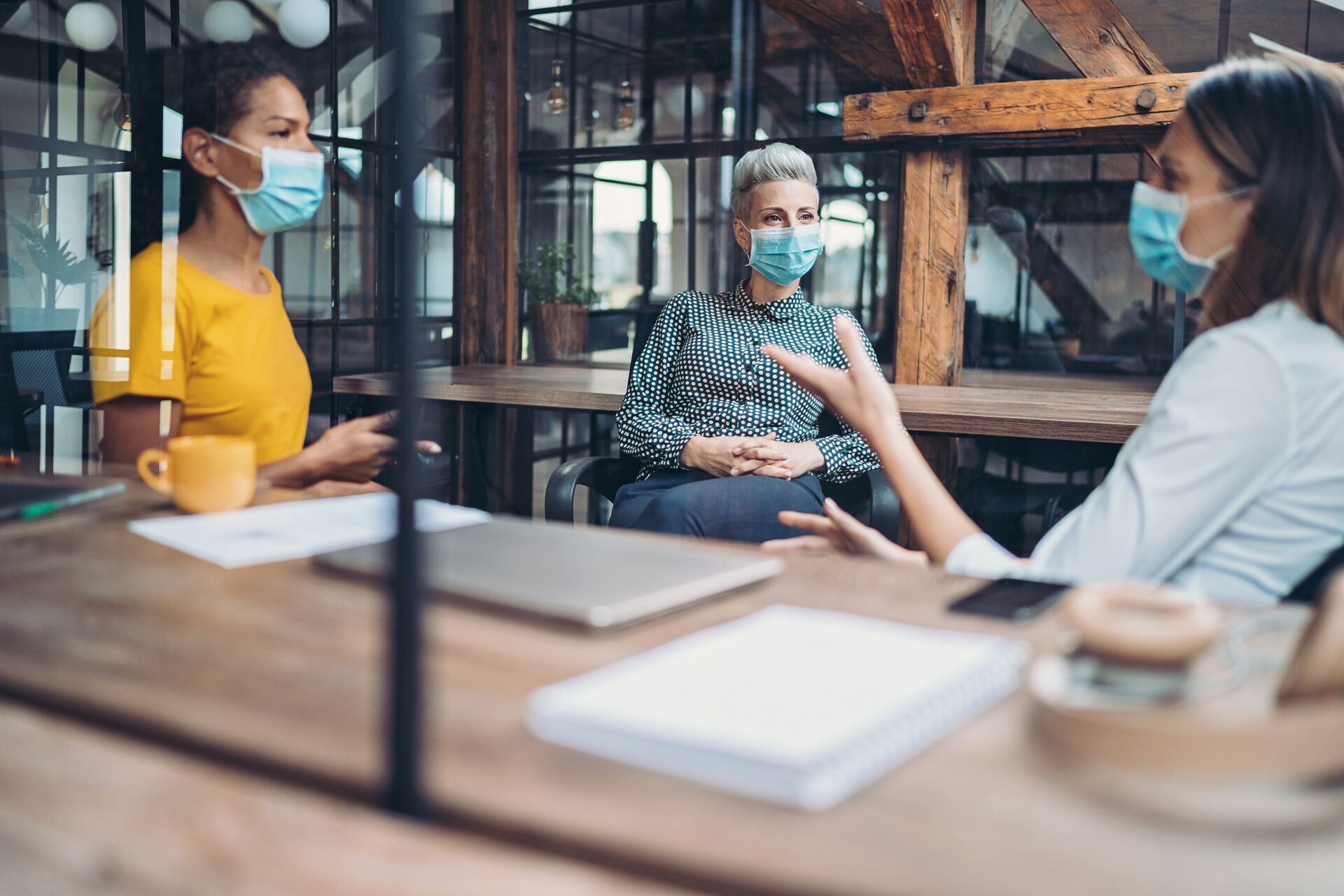 Three women sitting across table at work, discussing a project while wearing masks through a pandemic.